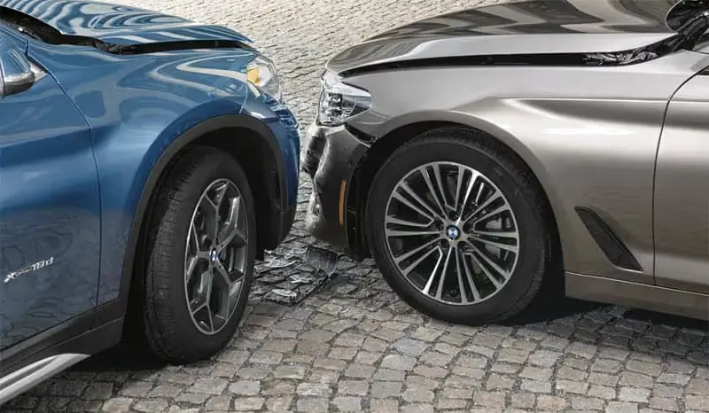 Front ends of two cars, a blue SUV and a tan sedan, with visible damage from a collision on a cobblestone road.