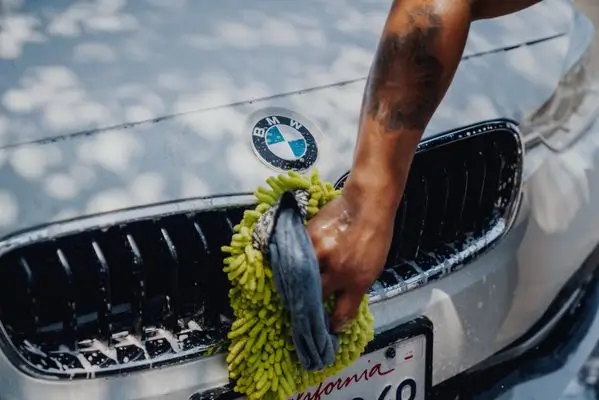 A dark-skinned hand with a tattoo holds a green chenille wash mitt, scrubbing white soap suds on a shiny gray BMW grille.