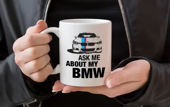 Hands holding a white mug with ASK ME ABOUT MY BMW and an illustration of a white BMW sedan with blue and red stripes.