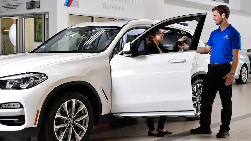 A salesperson hands car keys to a smiling woman in the driver’s seat of a white BMW SUV inside a dealership.